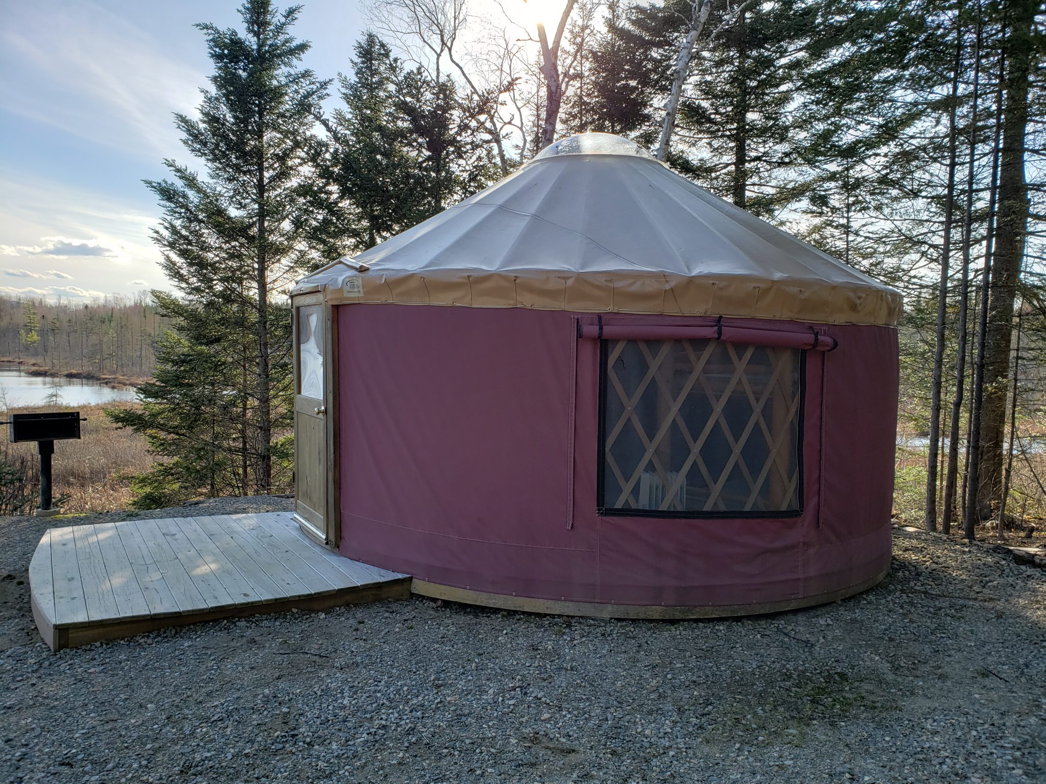 Maroon yurt with wooden deck and grill by a lake in a forest.