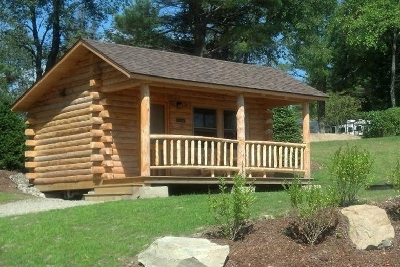Cozy log cabin with a wooden porch and railing in a grassy, wooded area.