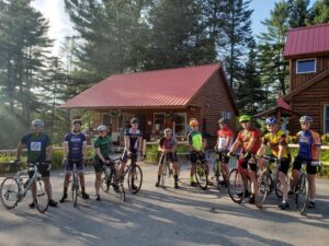 Group of cyclists in colorful gear posing with their bikes outside a rustic wooden building.
