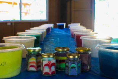 Jars of colorful layered sand art and large buckets of colored sand on a blue tablecloth.