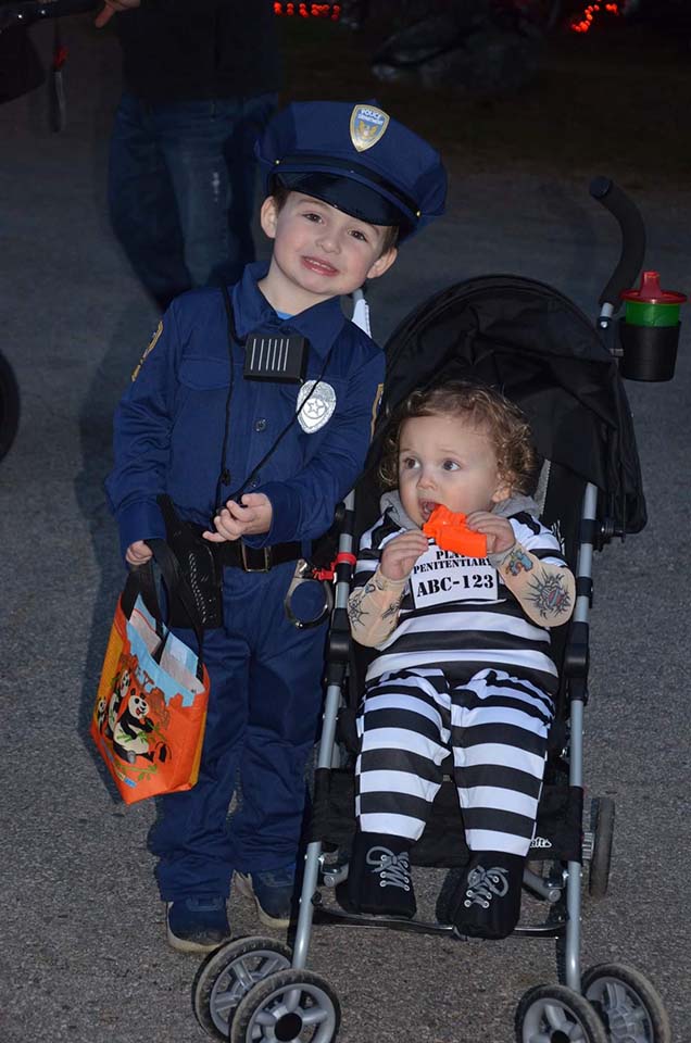 Boy in police costume with trick-or-treat bag, baby in prison costume in stroller.