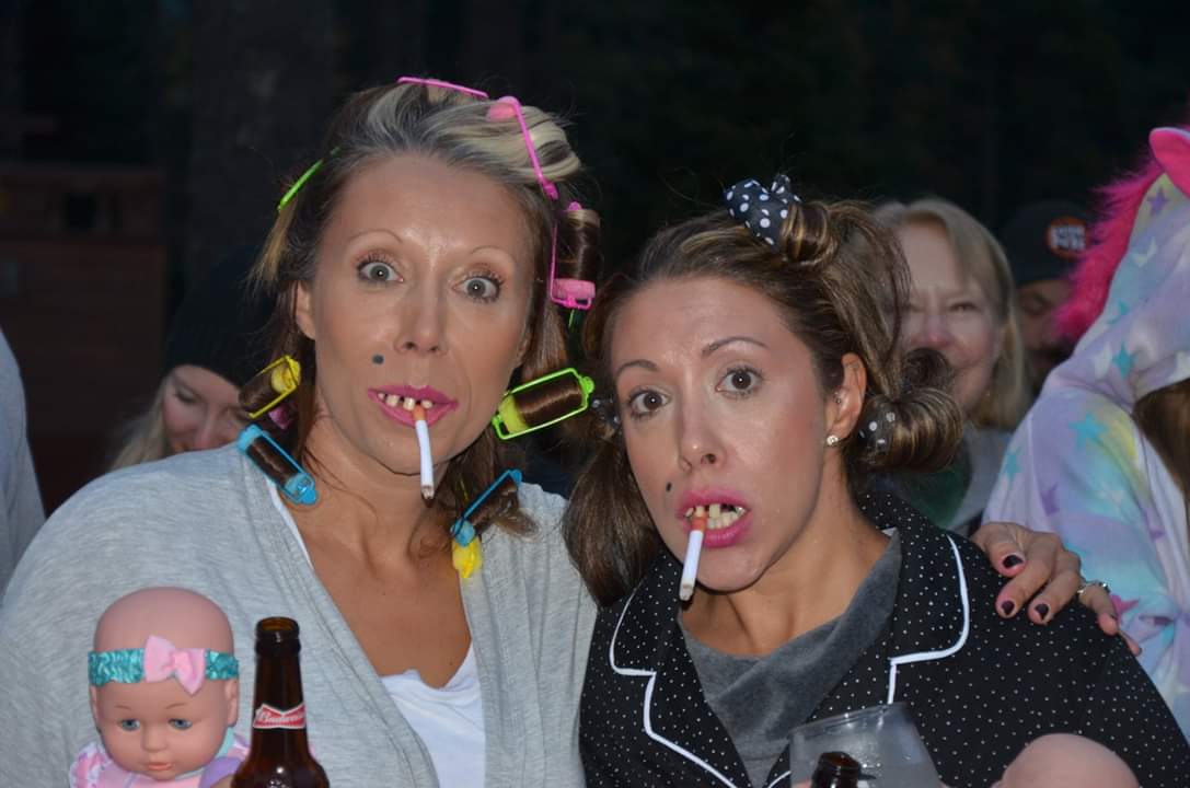 Two women in curlers and pajamas smoke cigarettes, dressed as "trailer trash" characters.