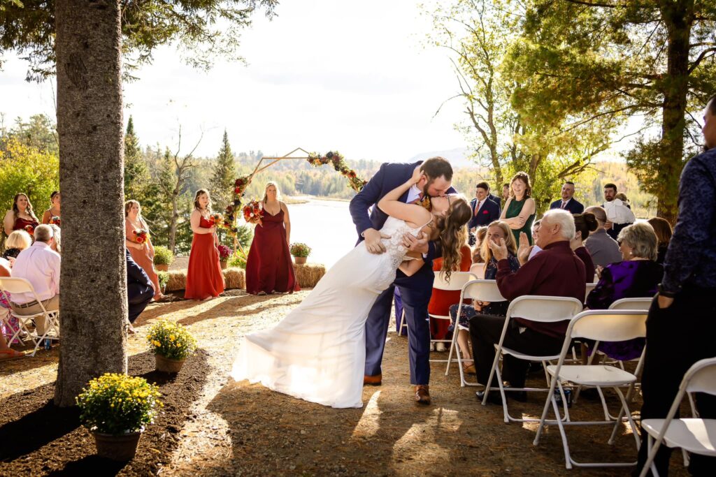 Bride and groom kiss at outdoor wedding ceremony with guests watching.